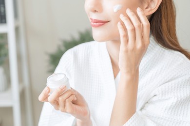 facial beauty skin care, smile of pretty asian young woman in bathrobe looking at mirror, hand applying moisturizer lotion on her face, holding jar of skin cream before makeup cosmetic routine at home