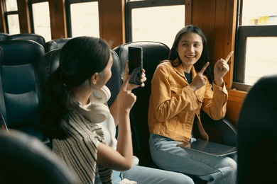 two female friends are sitting in a train, one is pointing at the window and the other is taking a picture with her phone