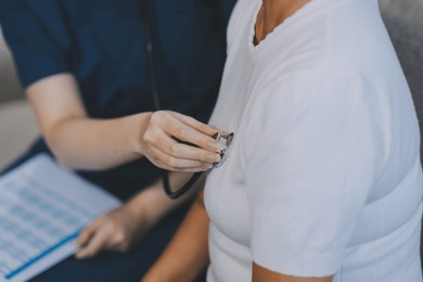 caucasian female doctor uses stethoscope to check heart rate on elderly asian patient on sofa.