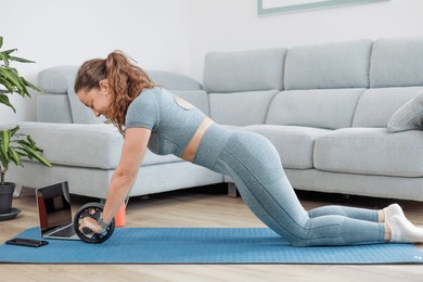 young athletic woman does sports with a roller or wheel on a mat in the living room at home with an online personal trainer
