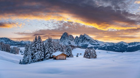 panoramic view on alpe di siusi meadow with cozy wooden log cabins and sunset sky. snowy hills with orange larch and first snow in winter season. seiser alm, dolomites, italy. landscape photography