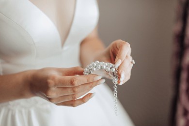 a woman is holding a necklace with pearls and diamonds. the necklace is white and has a gold chain. the woman is wearing a white dress and is standing in front of a curtain