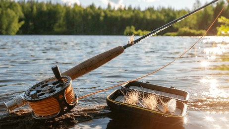 a fly fishing rod and an open fly fishing box lie on the rocks of a mountain river.