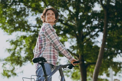 young smiling happy fun caucasian woman she wearing checkered shirt casual clothes look aside walk with bicycle rest relax in spring sunshine green city park outdoors on nature. urban leisure concept