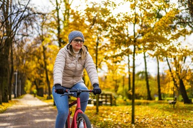 mid-adult blonde woman wearing beige jacket, jeans and winter hat riding bicycle in city park in autumnal scenery on sunny day. front view