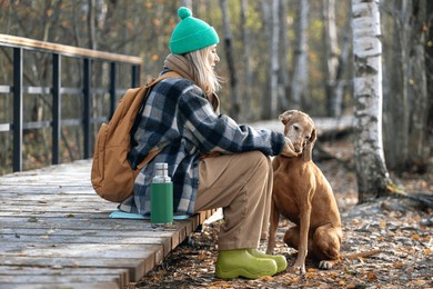 woman traveler pauses hike, feeds vizsla hunting dog by hand, sitting on eco trail in autumn nature parkland. female backpacker drinks tea, gives treats to pet, enjoys outdoor walk, active lifestyle
