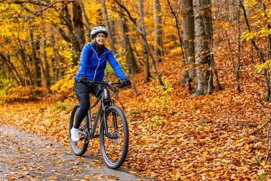 middle aged woman wearing blue sporty jacket, black cycling pants and white bike helmet riding bicycle in city forest in autumn scenery. front view.
