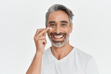 happy middle aged 45 years old man applying facial anti age skincare cream on face isolated on white background. smiling mature handsome gray-haired male model advertising skin care product. portrait.