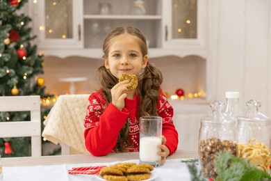 happy joyful cute little girl in red sweater drinking milk and eating delicious cookie near christmas tree in the kitchen for christmas morning. christmas time. christmas holidays