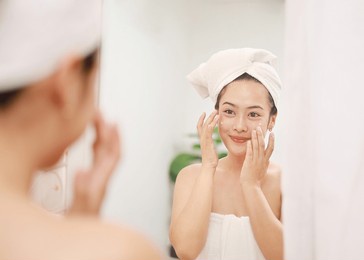 skin care beauty on face, smiling beautiful asian woman in bathrobe looking in mirror and using serum cream to nourish skin in daily life.