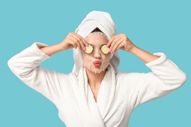 young woman in bathrobe with sheet facial mask and cucumber slices on color background