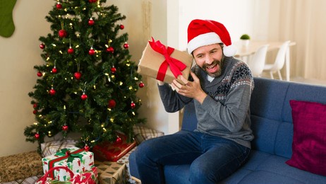 man eagerly shaking a christmas present and guessing its contents while seated on a festive sofa in a beautifully decorated living room