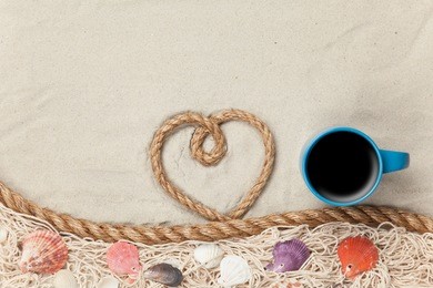 heart shape rope and cup of coffee near net and shells on sand.