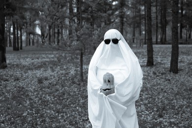 woman in white sheet holding jack o'lantern against autumn forest background. halloween costume. black and white photo. 