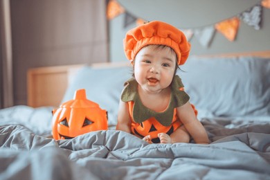 happy cute asian newborn baby girl dressed in halloween jack o lantern costume sitting on bed with halloween decoration, having fun and celebrating halloween at home