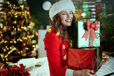 christmas time. happy elegant middle aged small business owner woman in santa hat and red jacket with present boxes in modern green office with christmas tree.
