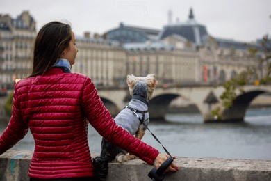 a beautiful young woman in red jacket with small yorkshire terrier dog sitting on parapet standing on the banks of a river seine in paris. journey to france with pet. walking outside on an autumn day.