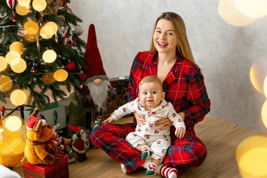 a joyful mother sits with her baby in pajamas next to a decorated christmas tree. the room is filled with festive decorations and gift-wrapped presents, creating a warm and cozy holiday atmosphere.