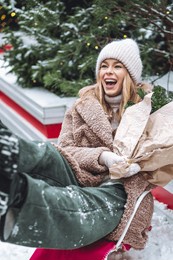 portrait of beautiful attractive smiling cheerful slavic young girl in a knitted hat and mittens with fir tree branches enjoying holiday christmas market, riding sled in the snow laughing. snowflakes 