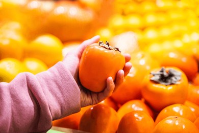woman holding fresh persimmon near fruit counter at market, closeup of female hand picking fresh persimmon at supermarket, grocery shopping concept