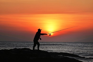 a person casting a fishing line against a vibrant sunset over the ocean.