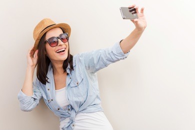 collecting memories. portrait of beautiful young woman in glasses adjusting her hat while making selfie and standing against brown background