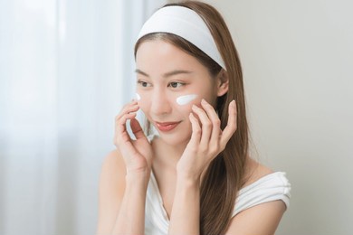 facial beauty, skin care concept, smile asian young woman applying moisturizer lotion on her face, pretty girl putting cream treatment before makeup cosmetic. isolated on background.
