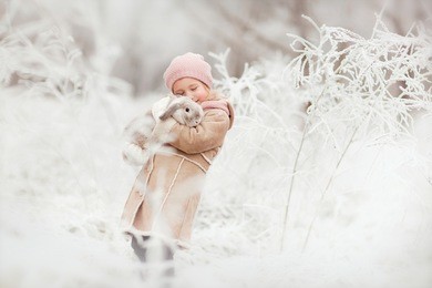 little cute smiling girl in pink hat and beige coat standing in the winter magic forest and holding a rabbit in her hands 