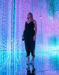 young woman stands surrounded by colorful lights in an immersive art installation.