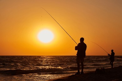 silhouettes of two people fishing on the beach at sunset, with the sun setting over the sea.