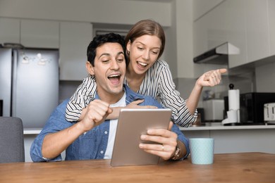 happy couple reading super news on digital tablet in kitchen, received great sell-out offer, commercial proposal, huge discounts or lottery win notice. moment of achievement, innovations, new software