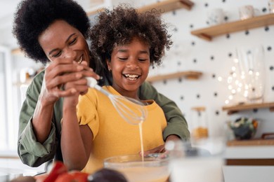 smiling african mother and daughter enjoy quality time cooking in a well-lit kitchen. the child holds a whisk, mixing ingredients while the mother lovingly guides her.