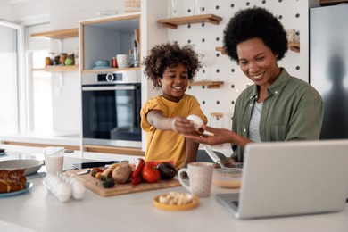natural light illuminates mid-aged african american mother and elementary-age son chopping vegetables together in contemporary white kitchen, sharing cooking moment near laptop.
