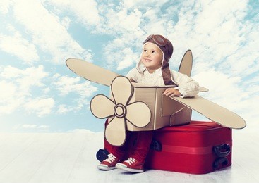 little child playing airplane pilot, kid traveler flying in aviator helmet on travel suitcase, vacation trip concept over blue sky clouds