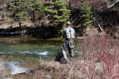 a man facing a flowing river and wearing fly fishing gear including a net hanging on his back fishes on the shore on a sunny day