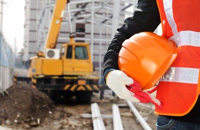 construction safety concept, close-up worker wearing safety vest holding helmet with crane in the background