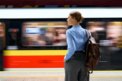 back view of young woman with brown leather backpack standing at subway platform, watching a fast-moving metro train pass by. urban commuting, city life, and public transport theme.