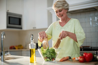 senior woman tossing green salad in home kitchen using glass bowl and wooden servers, copy space. stylish, wholesome, culinary, domestic, contemporary, vibrant, nourishing