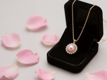 a beautiful pink pearl pendant necklace, set in gold with small sparkling stones, rests in a black velvet gift box. pink rose petals and a large rose adorn the background.