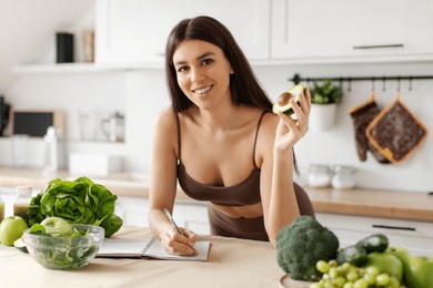 happy slim european woman planning daily meals, writing weight loss diet plan, holding avocado and smiling at camera in kitchen interior