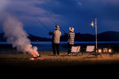 night fishing with friends by lake under lantern light, camping and fishing near campfire at twilight under blue sky, scenic night escape with fishing gear and fire, weekend lakeside activity
