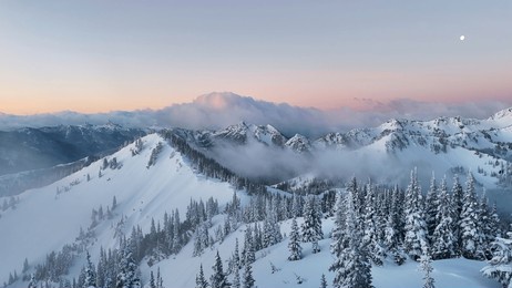 a breathtaking aerial view of snow covered mountains, trees, and clouds under a pastel sky at twilight