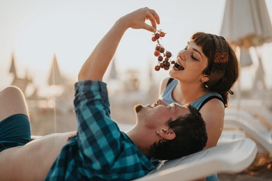 a young couple enjoying their vacation at a sunny beach, sharing grapes and relaxing on sun loungers with a joyful atmosphere under umbrellas.