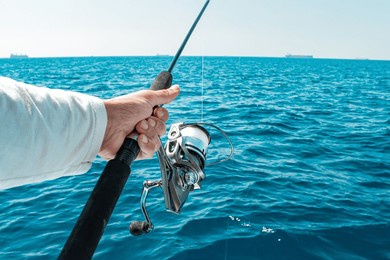 a fisherman hand firmly grips a fishing rod with a modern silver spinning reel ready to cast into the bright blue sea where distant cargo ships dot the horizon
