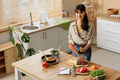 young woman with recipe book grating carrot on table in kitchen