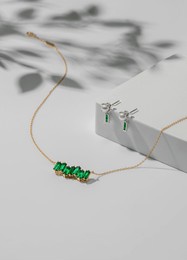 a lovely necklace featuring emerald green stones is paired with matching earrings, elegantly arranged on a white pedestal. soft lighting enhances the jewelry's shine.