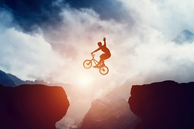 man jumping on bmx bike over precipice in mountains at sunset. raising hand showing hello gesture. extreme sport, risk, cycling.
