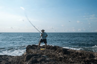 angler fishing from rocky coastline on sunny day with clear blue sky and ocean waves - outdoor recreation and leisure activity by the sea