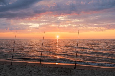 three fishing rods stand on a sandy beach, silhouetted against a breathtaking and colorful sunrise over the tranquil ocean.