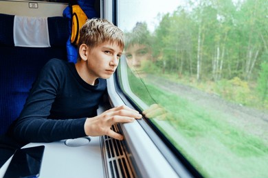 teen boy pressing head against window while riding train, observing outside landscape, hands on sill, serious expression, solo travel, forest visible through glass